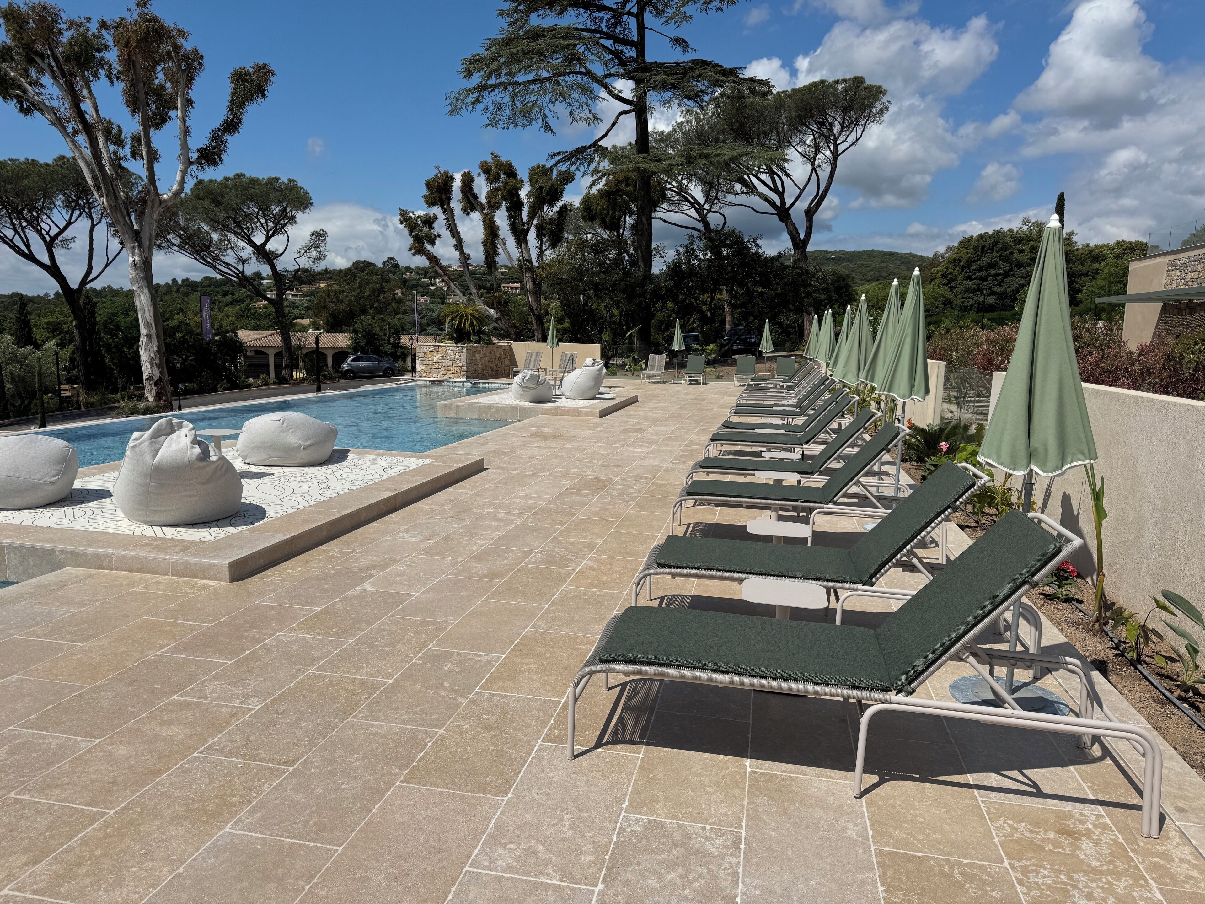 A poolside area with lounge chairs and umbrellas, surrounded by trees and a clear sky.