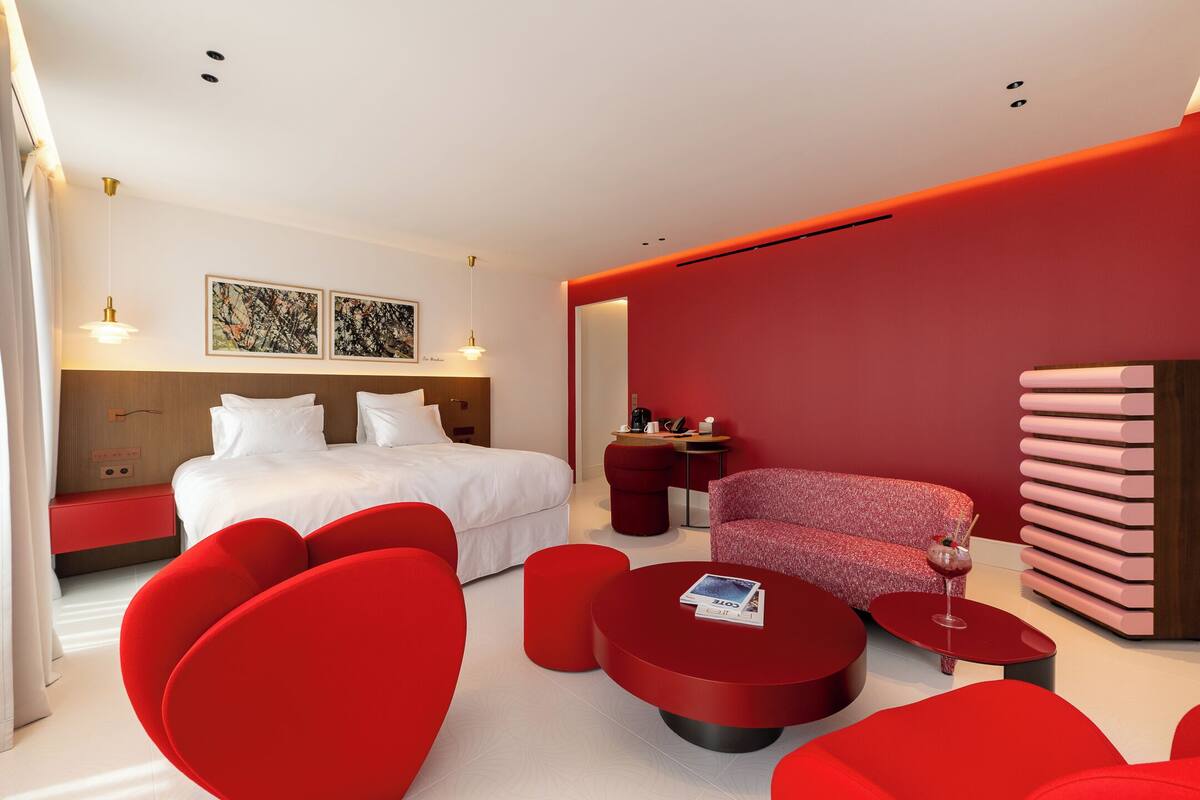 Ein modernes Hotelzimmer mit Bett, roten Möbeln, einem kleinen Tisch mit einem Getränk und Kunstwerken an der Wand.