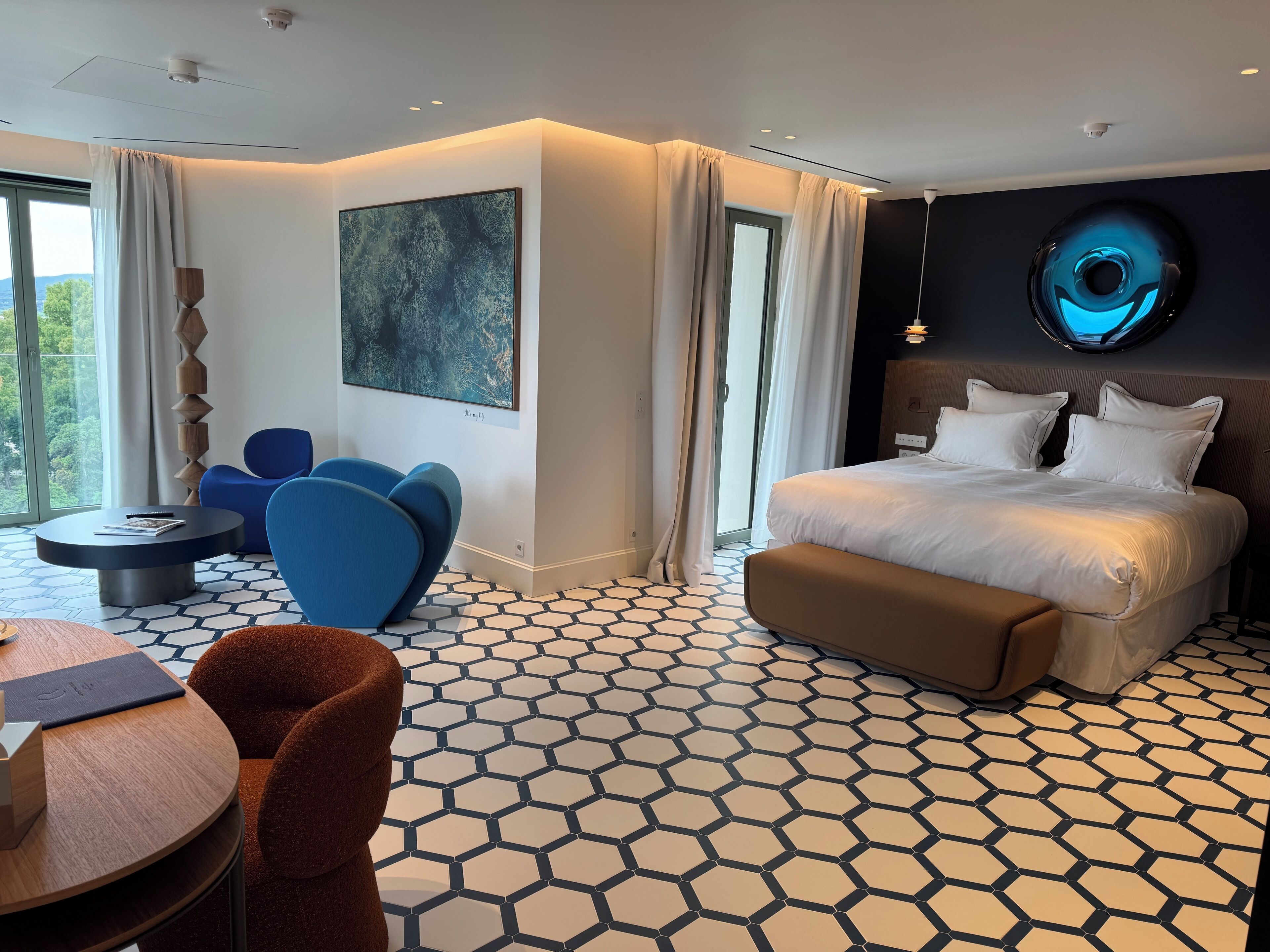 A modern hotel room with a large bed, a round dining table, and a seating area with blue chairs.