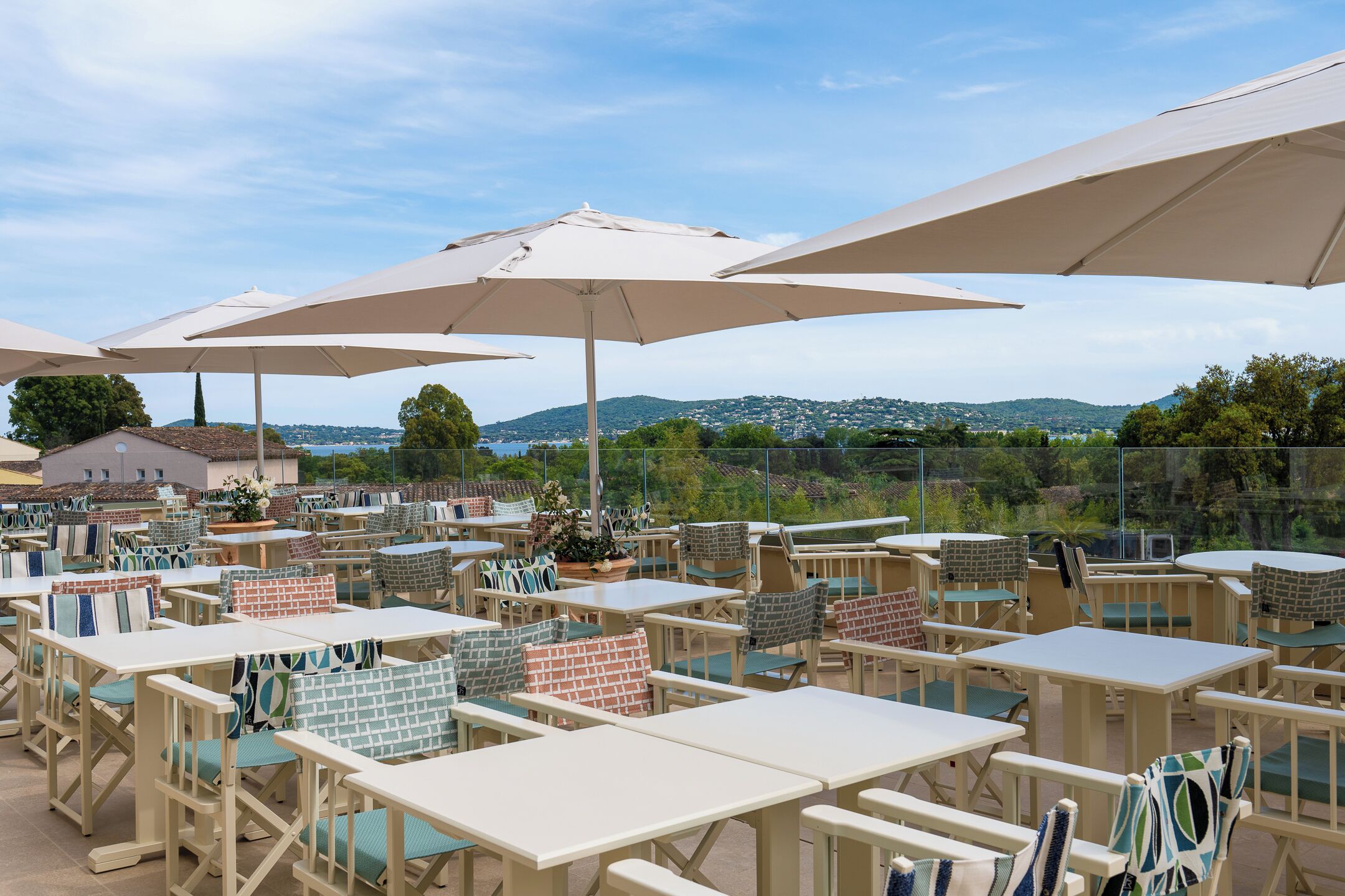 Un patio al aire libre con varias mesas y sillas, grandes sombrillas y una vista panorámica de colinas y árboles.
