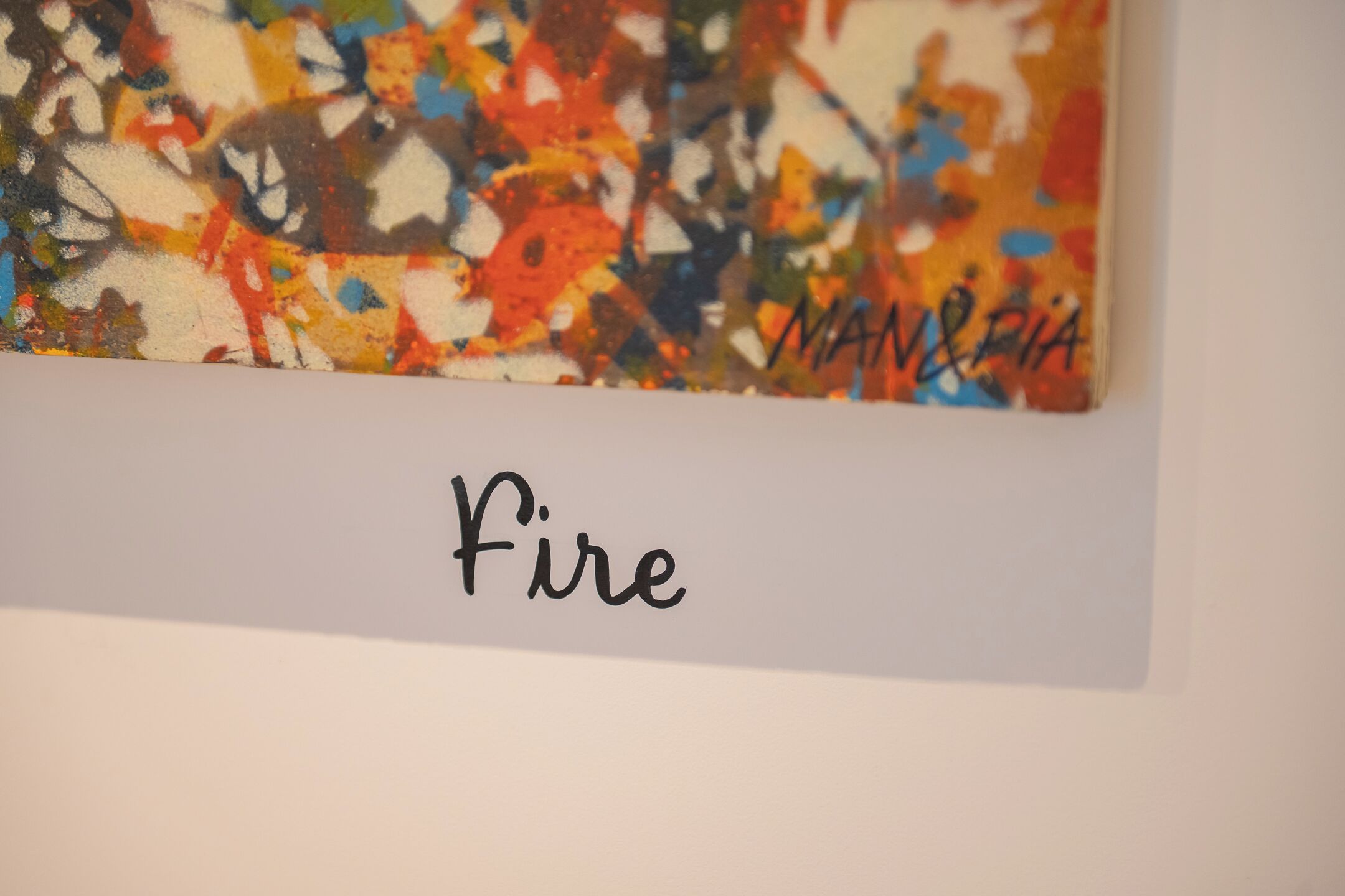 Una pintura abstracta y colorida con la palabra “Fire” y la firma del artista “MAN & RIA” sobre un fondo blanco.