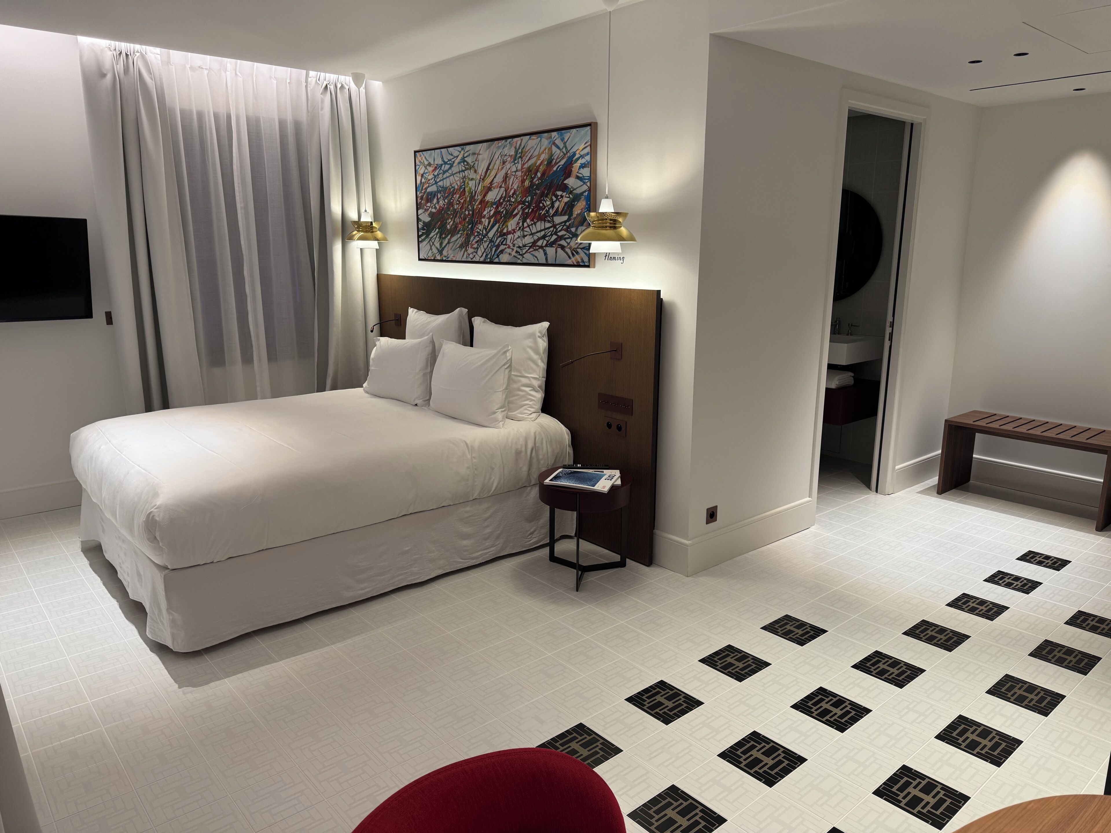 A modern hotel room with a large bed, a bedside table, a bench, and a painting on the wall.