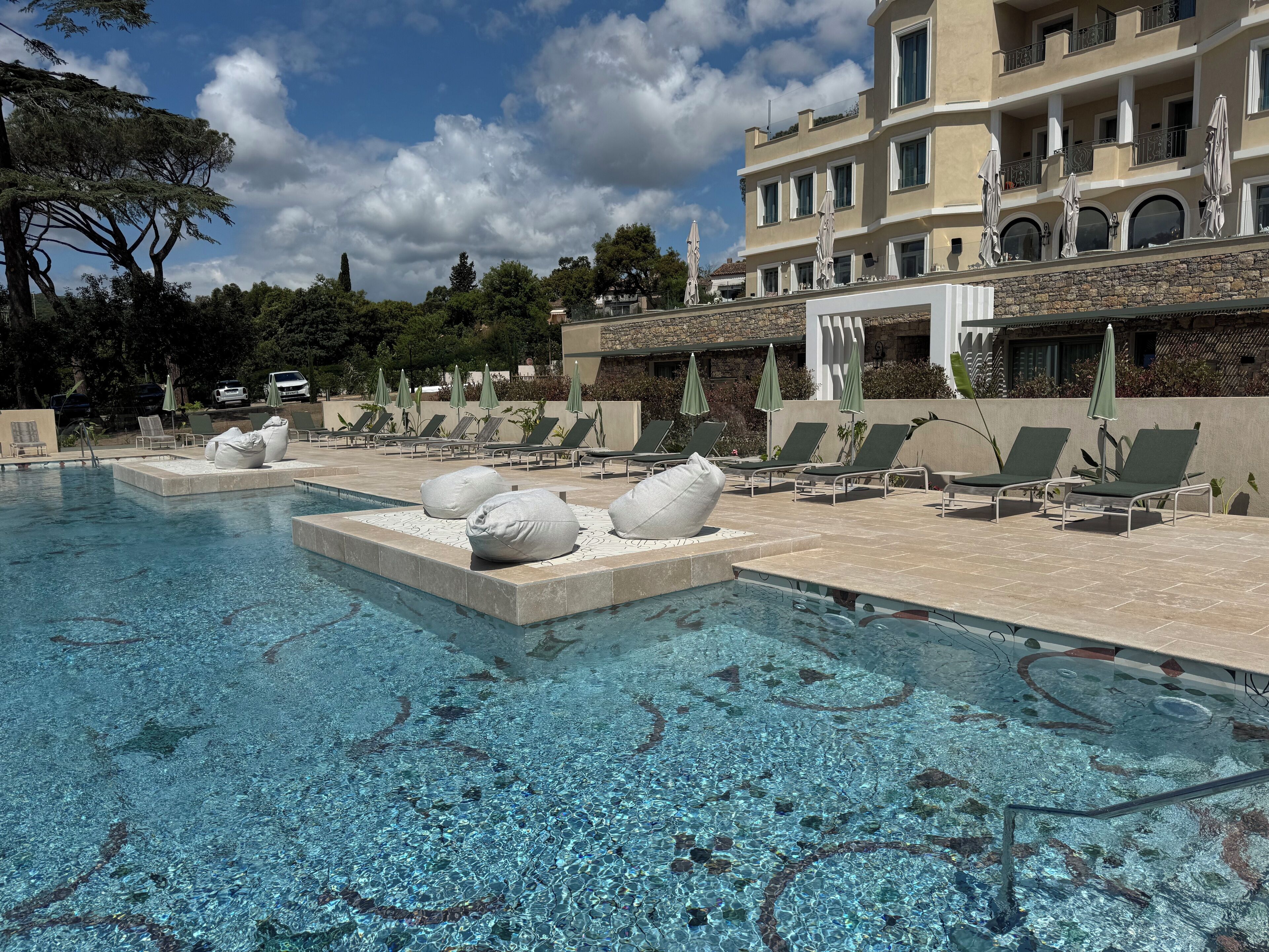 Un área de piscina con sillones reclinables y sombrillas, una piscina cristalina y un edificio con balcones.