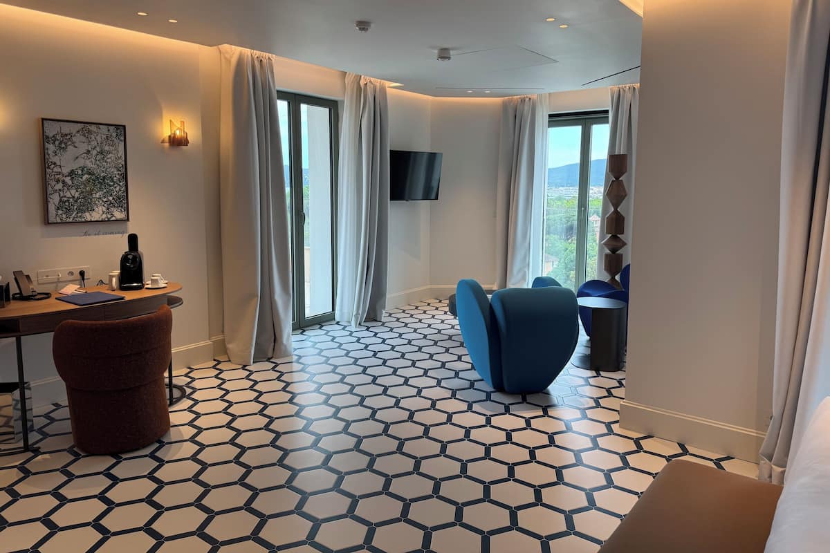 Ein modernes Hotelzimmer mit einem sechseckig gemusterten Boden, einem Schreibtisch mit Kaffeemaschine, einem braunen Hocker, einem blauen Sessel und einem an der Wand montierten Fernseher.