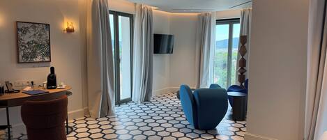 Una habitación de hotel moderna con piso de patrón hexagonal, un escritorio con cafetera, un reposapiés marrón, una silla azul y un televisor montado en la pared.