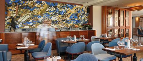 Un restaurant avec des tables rondes, des chaises bleues et un grand tableau abstrait accroché au mur.