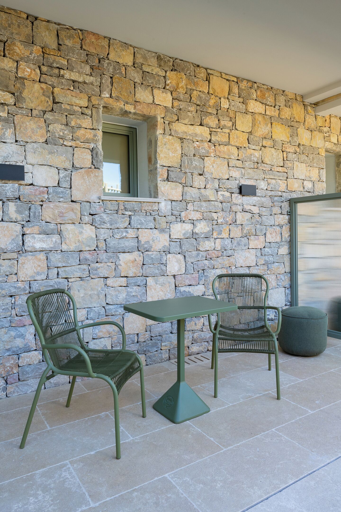 A patio with two green metal chairs and a table, set against a stone wall with a window and a door.