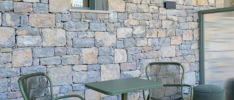 Un patio con dos sillas metálicas verdes y una mesa, frente a un muro de piedra con una ventana y una puerta.