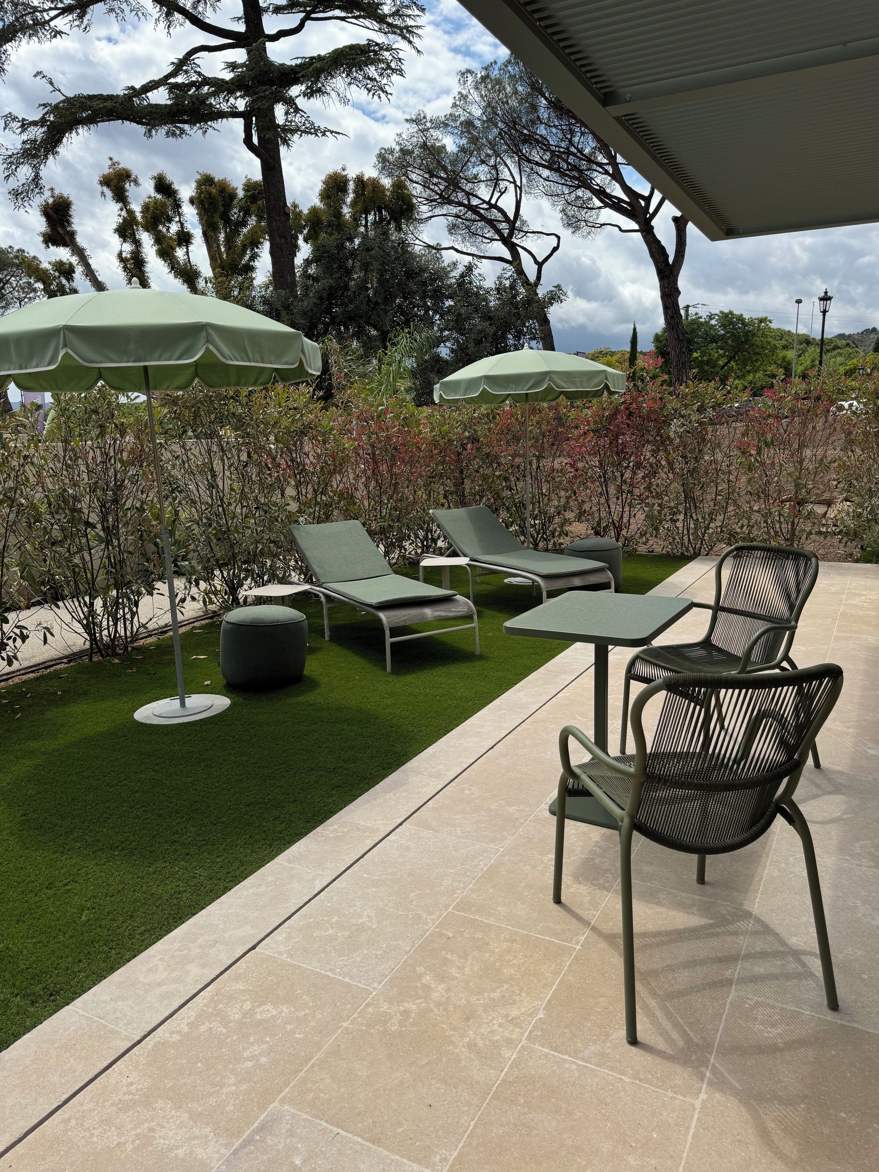 Un patio con césped artificial verde, sillas de descanso y mesas bajo sombrillas verdes.