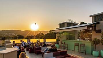 Un bar en la azotea con mesas al aire libre, zona de bar y vistas a la puesta de sol sobre un paisaje montañoso.