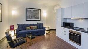 Ett modernt vardagsrum med en blå soffa, svarta läderfåtöljer och ett litet bord. Ett inramat konstverk hänger på väggen, och ett kök med vita skåp och vitvaror i rostfritt stål syns.