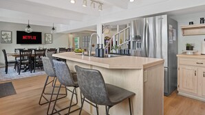 Une cuisine moderne dotée d’un îlot central, de tabourets de bar, d’un réfrigérateur en acier inoxydable et d’un coin repas avec un téléviseur diffusant du contenu « NETFLIX ».