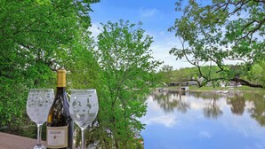 Une bouteille de vin et deux verres sont posés sur une étagère en bois, surplombant un lac paisible, avec des arbres et un pont en arrière-plan.