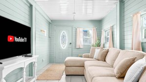 A living room with a beige sofa, a white console table with a flat-screen TV displaying the YouTube TV logo, a door with a decorative glass panel, and a window with curtains. - Walk to Vilano Beach + Pool + Hot Tub + Pets (St. Augustine)