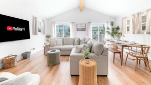 A modern living room with a large flat-screen TV, a grey sectional sofa, a dining table with chairs, and a large window with curtains. - Walk to Vilano Beach + Pool + Hot Tub + Pets (St. Augustine)
