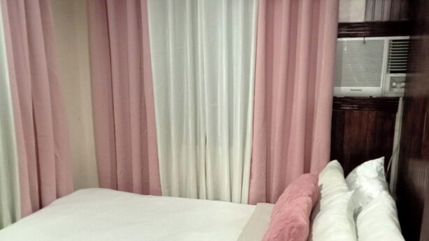 Ein Einzelbett mit weißer Matratze und rosa Bettwäsche, ein hölzernes Kopfteil sowie ein Fenster mit rosa-weißen Vorhängen.