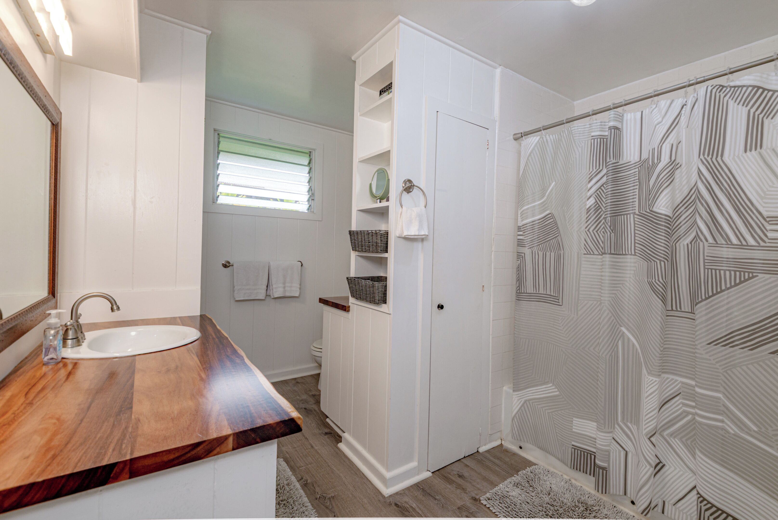 A bathroom with a wooden vanity, a white sink, a mirror, a towel rack, a shower with a patterned door, and a window with blinds.
