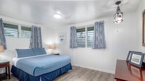 A bedroom with a bed, a nightstand, a lamp, a window with curtains, a ceiling fan, and a picture frame on the wall. - Kai Cottage - A Little Piece of Old Hilo (Hilo)