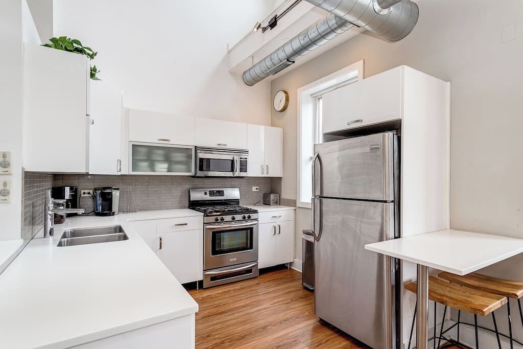 Une cuisine moderne avec des meubles blancs, des appareils en acier inoxydable, un plancher en bois et une petite table à manger.