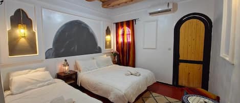 A room with two beds, a small table, a colorful rug, a wooden door, and a ceiling fan.