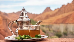 Een traditionele metalen theepot en glas thee, geplaatst op een bord met muntblaadjes, met een bergachtige achtergrond.