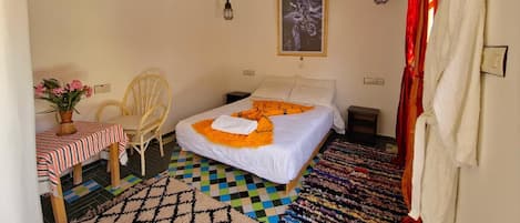 A bedroom with a bed, a bedside table, a chair, a small table with a vase of flowers, a patterned rug, and a ceiling fan.