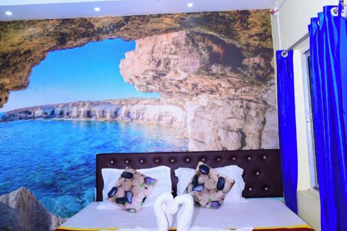 A hotel room with a bed, a ceiling fan, a wall mural of a coastal scene, and blue curtains.