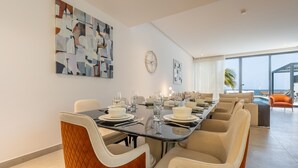 A dining area with a long table set for six, surrounded by cushioned chairs, a large abstract painting on the wall, and a view of the ocean through large windows. - Exclusive 4BR Villa, Lush Garden & Private Pool Facing Panoramic Sea Views (Fujairah)