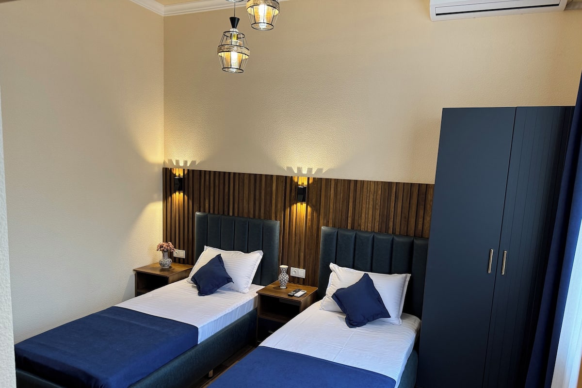 A hotel room with two beds, wooden headboards, bedside tables with lamps, and a wardrobe.