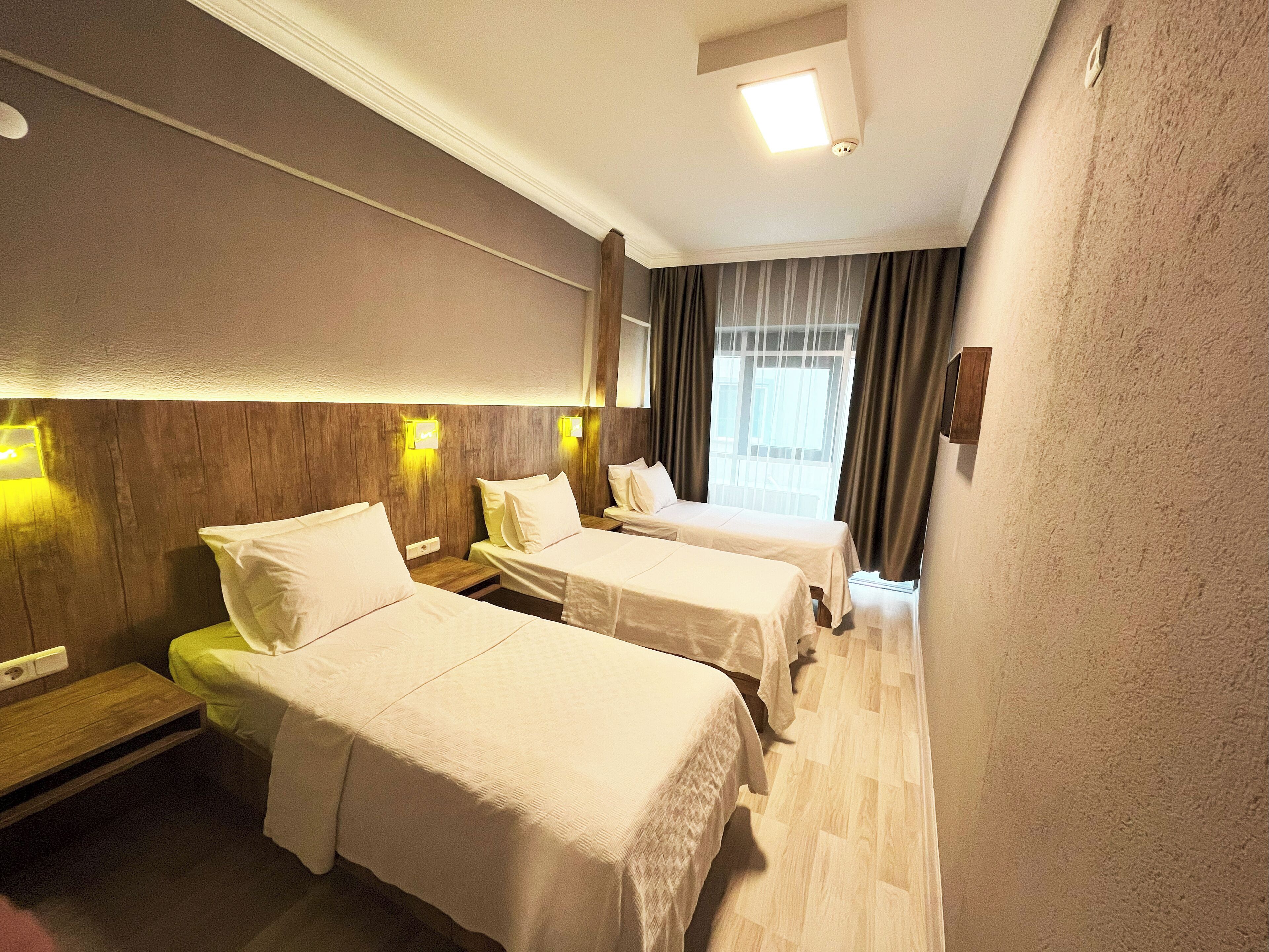 Una habitación de hotel con dos camas, cabeceros de madera, una mesita pequeña y una ventana con cortinas.