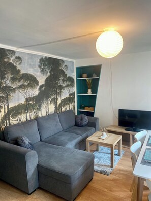Un salon moderne avec un canapé d’angle gris, une petite table basse en bois, un téléviseur à écran plat et une étagère turquoise.