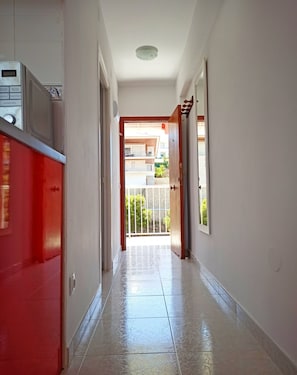 Um corredor com uma porta vermelha à esquerda, uma porta branca à direita e vista para uma varanda com guarda-corpo.
