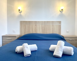 Cama bem arrumada com colcha azul e duas toalhas brancas dobradas sobre ela. Há duas mesas de cabeceira com abajures de cada lado da cama.