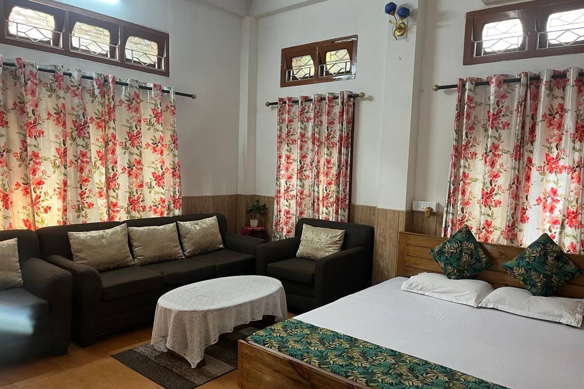 A room with a bed, two sofas, a small table, and floral curtains.
