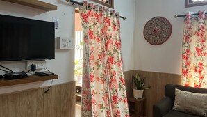 Un salon doté d’un téléviseur à écran plat, d’étagères en bois, d’une plante en pot, d’un élément décoratif rond fixé au mur et de rideaux à motifs floraux.