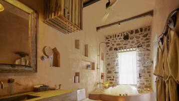 A rustic bathroom with a stone wall, a large bathtub, a wooden ceiling, and a mirror with a wooden frame.