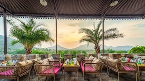 A covered outdoor seating area with wicker furniture and a table set for dining, overlooking a mountainous landscape. - The Quila Udaipur by Aayor Luxury (Mavli)