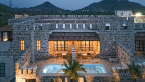 A stone building with a swimming pool, outdoor seating, and a view of mountains. - The Quila Udaipur by Aayor Luxury (Mavli)