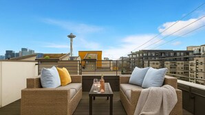 A rooftop patio with wicker furniture, a small table with glasses, and a view of the city skyline including the Space Needle.