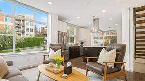 A modern kitchen with a large window, a refrigerator, and a dining area with two leather chairs and a wooden coffee table.