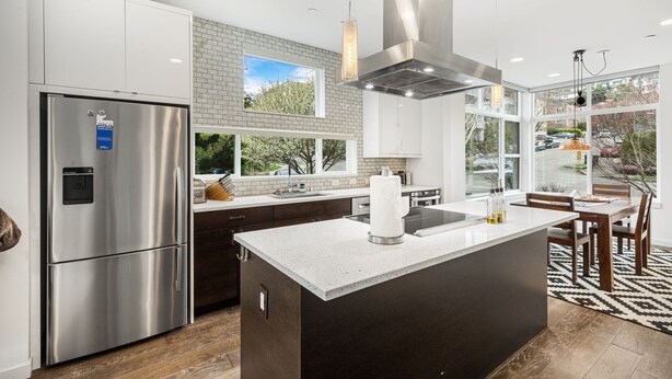 A modern kitchen with a stainless steel refrigerator, a large island with a sink, and a view of the outdoors through large windows.