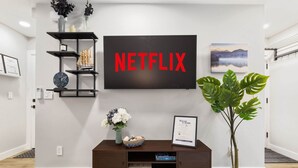 A modern living room with a mounted TV displaying "NETFLIX," a dark wooden console table with decorative items, a potted plant, and a framed welcome sign. - Sauna/Gym | Near Beach & Ballard | Dogs OK (Seattle)