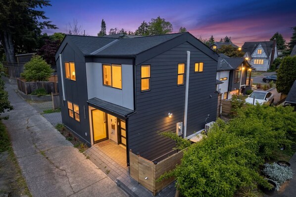 A modern two-story house with a dark exterior, multiple windows, and a well-lit entrance.