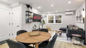 A modern living room with a round wooden table, grey upholstered chairs, a grey sofa, and a small coffee table. There is a bookshelf with decorative items and a wall-mounted TV displaying the Netflix logo. - Sauna/Gym | Near Beach & Ballard | Dogs OK (Seattle)