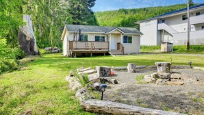 A two-story cabin with a deck, surrounded by a grassy yard and a forested hillside.