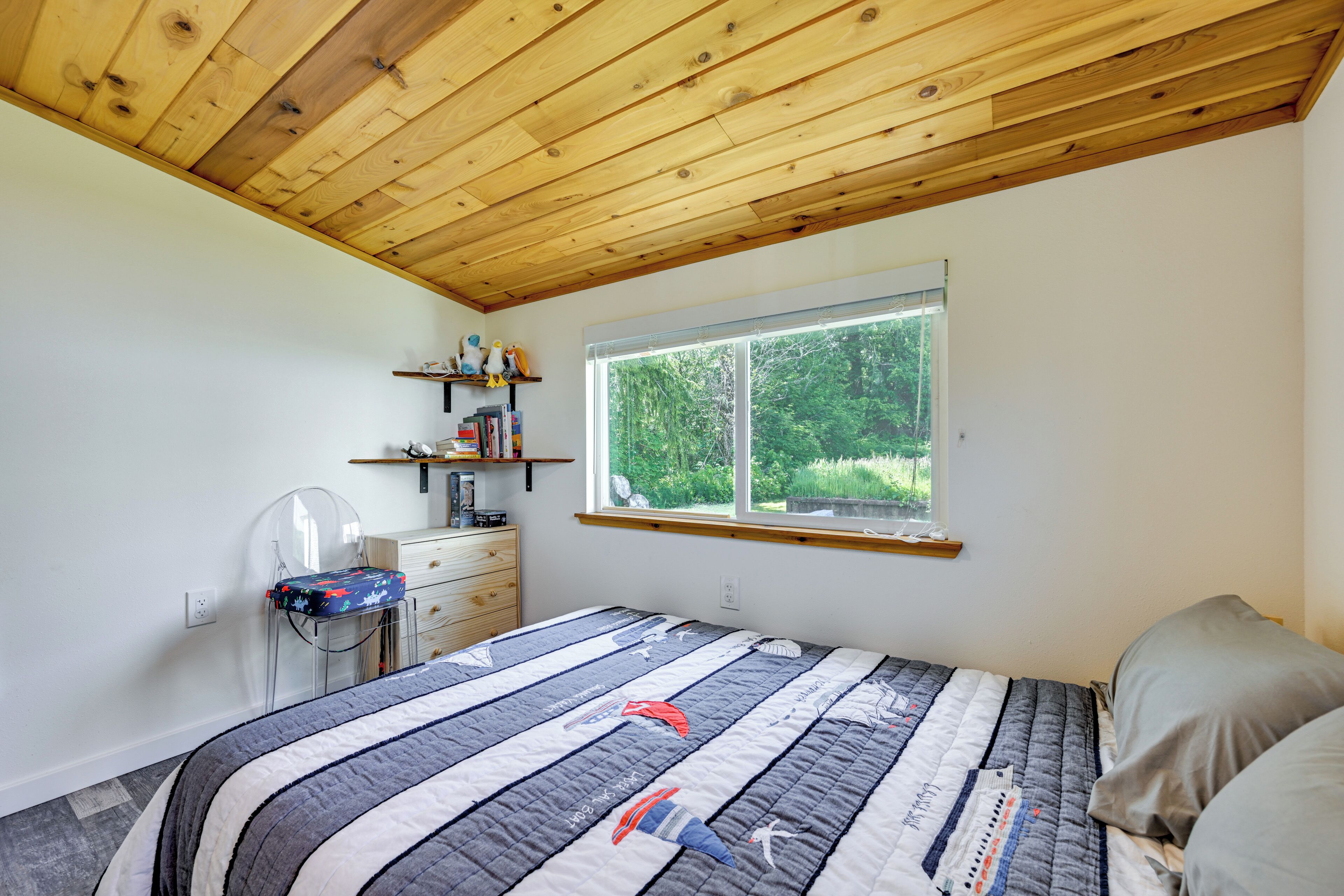 A bedroom with a wooden ceiling, a large window with a view of greenery, a bed with a striped quilt, a desk with a chair, and a shelf with books and decorative items.