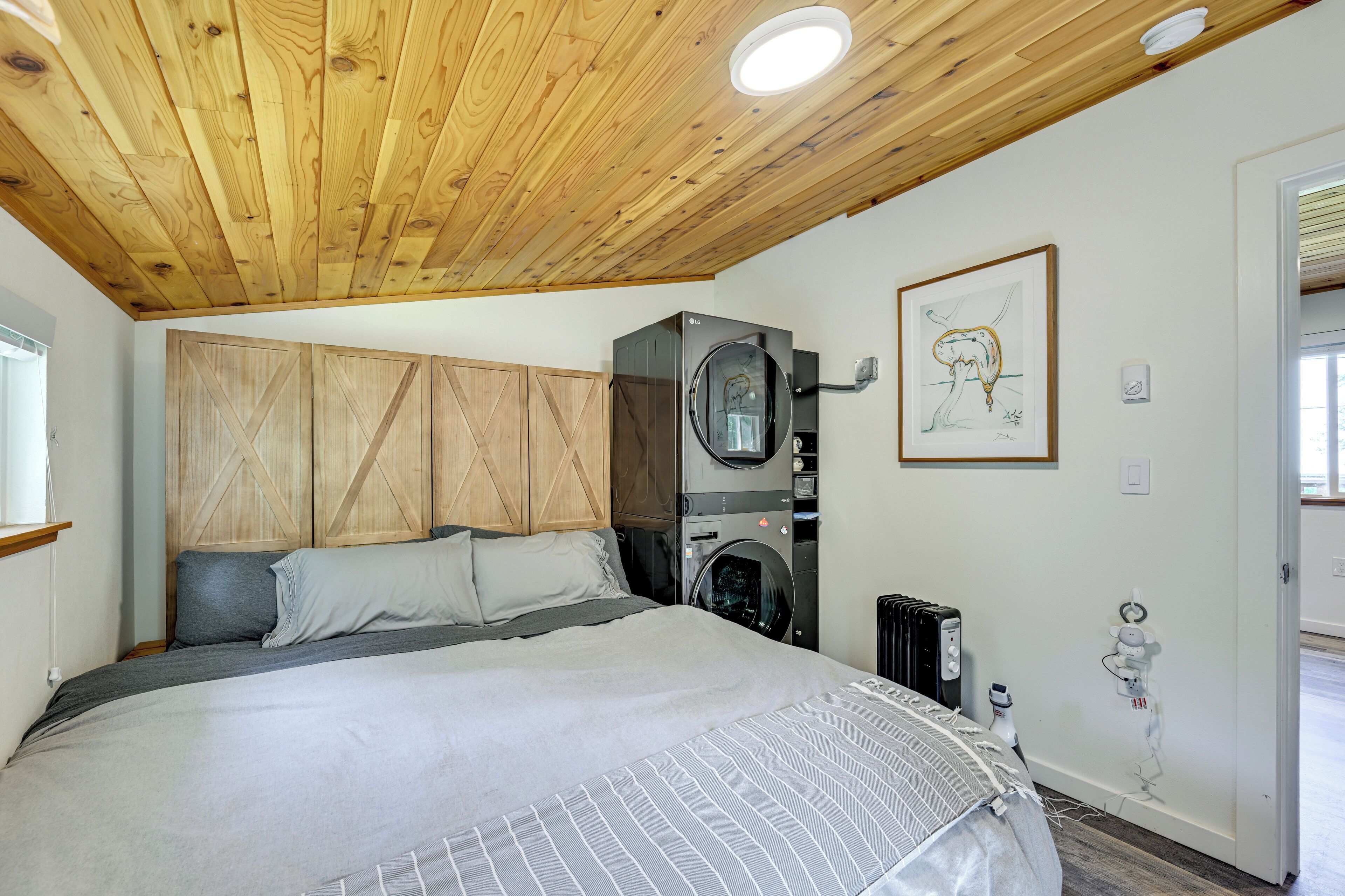 A bedroom with a wooden ceiling, a bed with grey bedding, a washing machine, and a radiator.