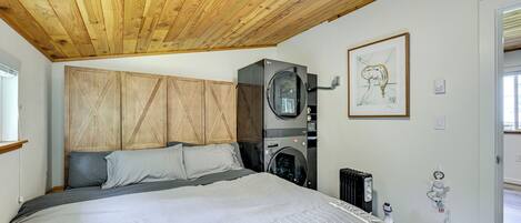 A bedroom with a wooden ceiling, a bed with grey bedding, a washing machine, and a radiator.