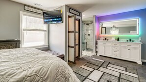 Une chambre à coucher avec un lit, un téléviseur fixé au mur, une salle de bain avec un grand miroir et une douche.