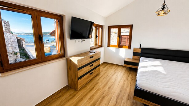 Un dormitorio con un aparador de madera, una cama, un televisor montado en la pared y ventanas grandes con vista al mar.